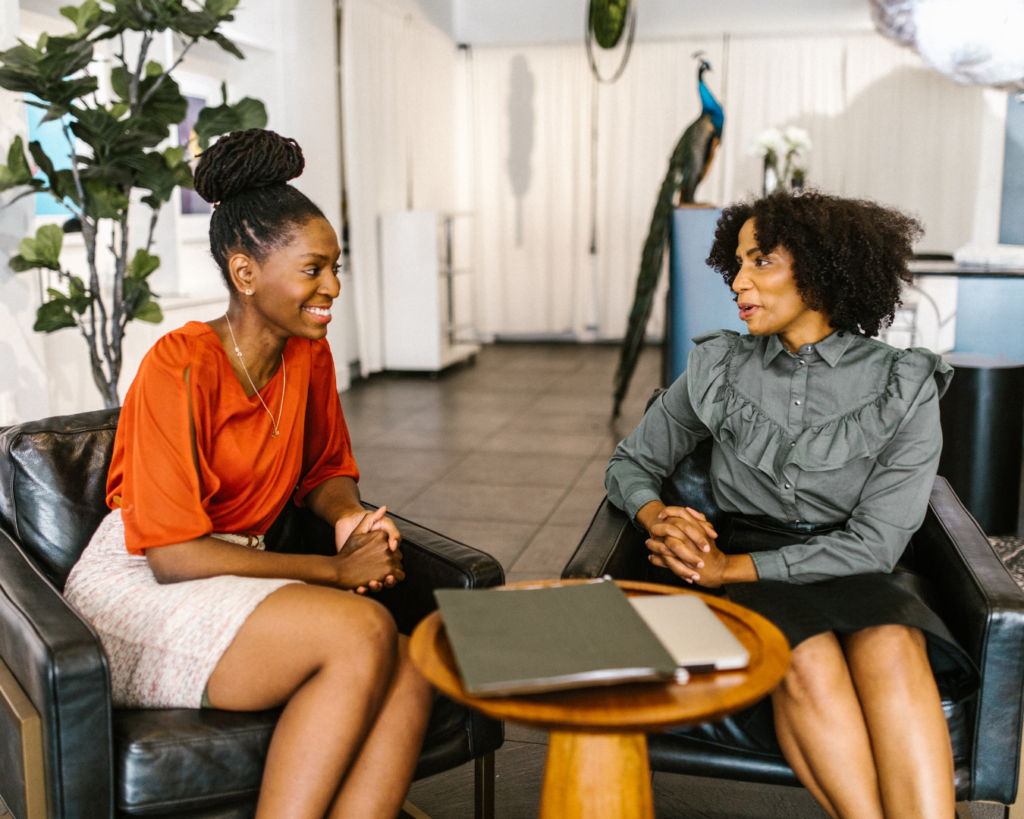 Two women in a coaching-style conversation on lounge chairs with a notebook on the table, reflecting on career clarity questions for midlife work decisions.