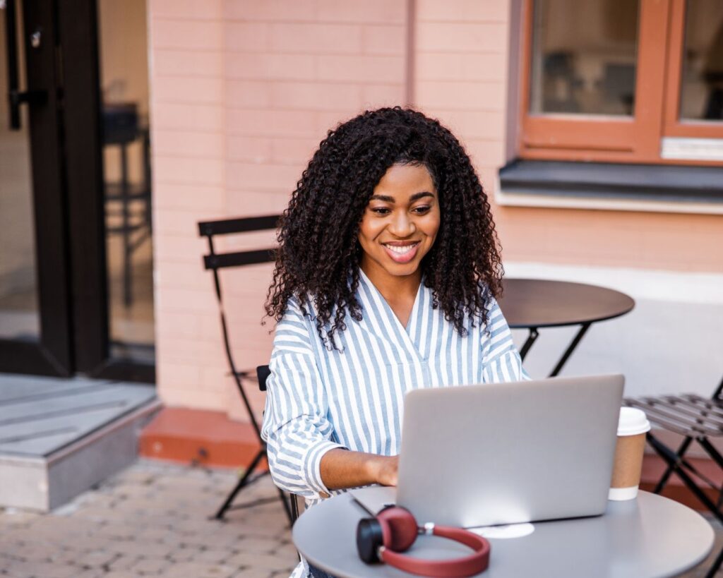 Smiling woman working on a laptop at an outdoor café table with a takeaway coffee, representing a midlife boundary reset and healthier work routines.