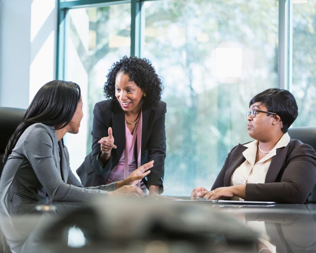 Three women in a workplace meeting, one standing and speaking while two listen, representing midlife leadership and the midlife career advantage.