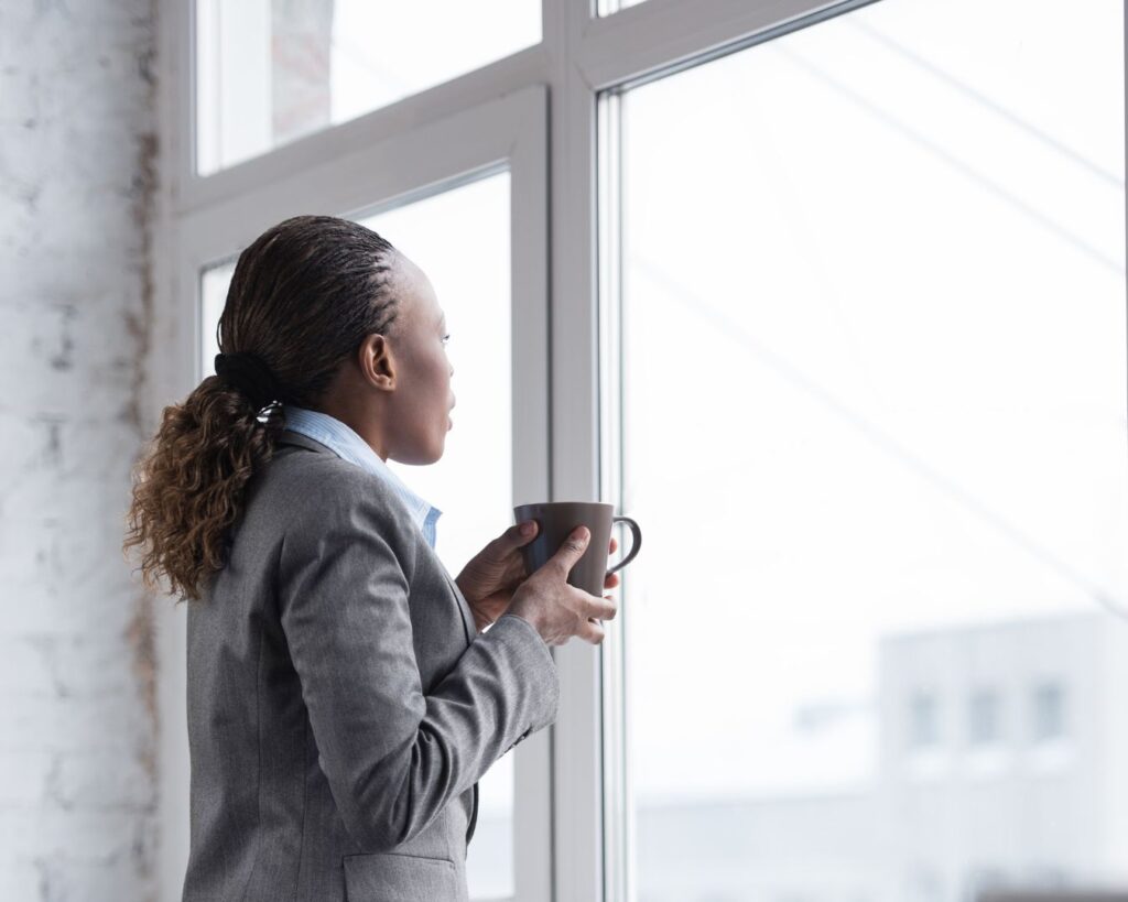 Black woman in a blazer stands by a large window holding a mug, reflecting quietly during a moment of career clarity and transition in midlife.